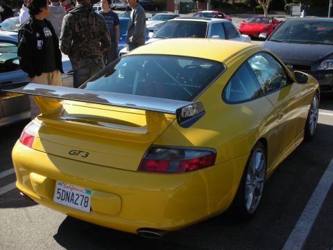 1998 Ford Crown Victoria - rear of a yellow Porsche 911 GT3 with a fixed rear wing and GT3 badge, parked at a car meet with spectators behind it.