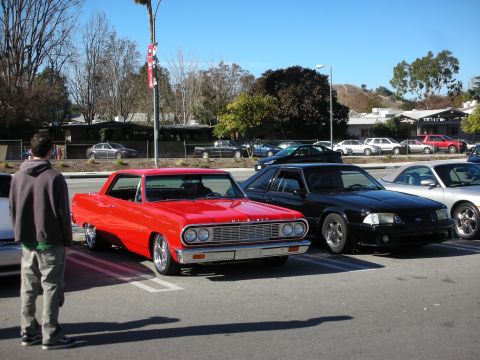 1998 Ford Crown Victoria - red 1960s Chevrolet Chevelle and a black Fox-body Ford Mustang park nose-out in a lot as a bystander looks on.