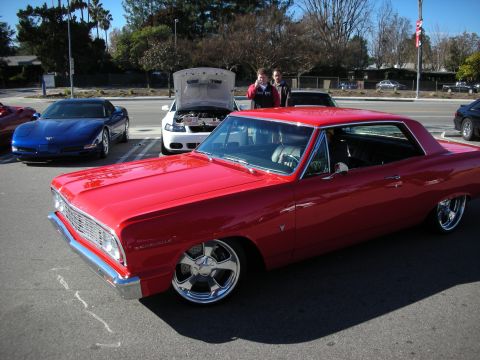 1998 Ford Crown Victoria - red 1960s Chevrolet Chevelle coupe on chrome wheels sits at an outdoor car gathering with two people standing behind it.