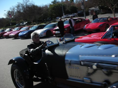 1998 Ford Crown Victoria - gray-haired man in a leather jacket drives a polished silver vintage roadster past a row of parked supercars at a meet.