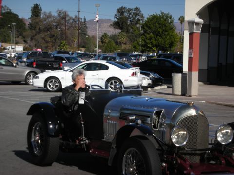 1998 Ford Crown Victoria - man pilots a riveted silver vintage roadster through a parking lot, with a white Ford Mustang and hills in the background.
