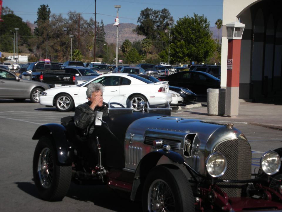 Man driving a silver vintage roadster past a white Ford Mustang in a parking lot