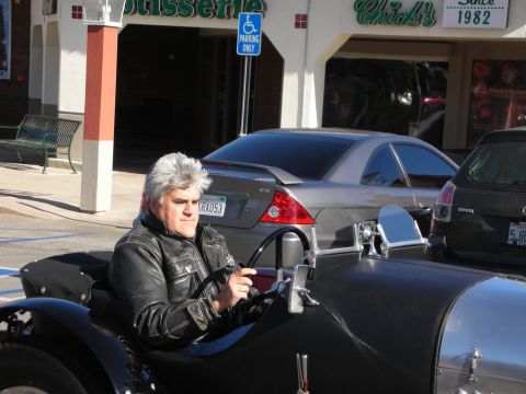 1998 Ford Crown Victoria - gray-haired man in a leather jacket sits in a vintage open-cockpit roadster on a street outside a storefront reading Chick's since 1982.