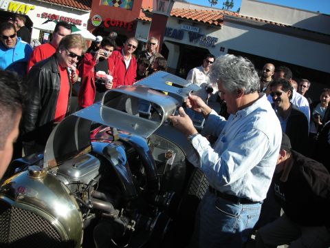 1998 Ford Crown Victoria - gray-haired man in a denim shirt lifts the metal hood panel of a vintage roadster while a crowd watches at a storefront.