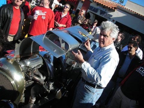 1998 Ford Crown Victoria - gray-haired man gestures beside the raised hood and engine of a vintage car as a crowd in red shirts looks on.