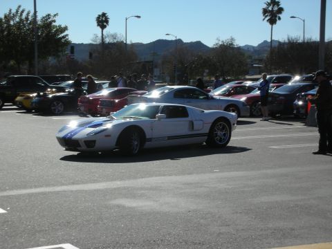 1998 Ford Crown Victoria - white Ford GT with twin blue racing stripes cruises through a sunny parking lot lined with cars and spectators.