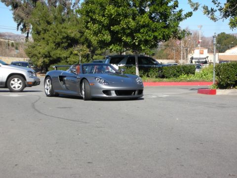 1998 Ford Crown Victoria - silver-gray Porsche Carrera GT roadster maneuvering through a parking area near trees and a fast-food sign.