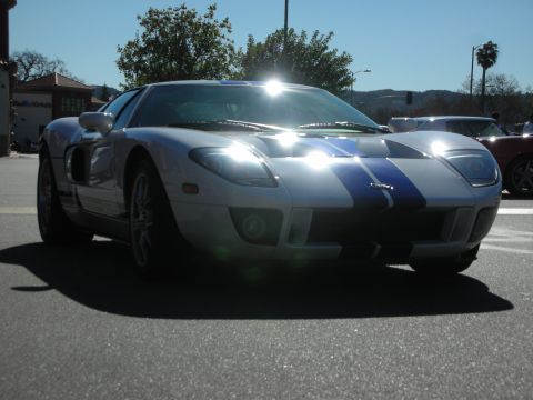 1998 Ford Crown Victoria - silver Ford GT with blue centre stripes and its headlights illuminated, parked at an outdoor gathering.