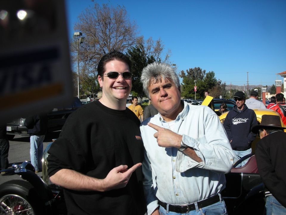 Two smiling men posing together at an outdoor car show, one with gray hair