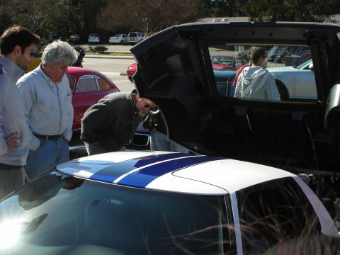 1998 Ford Crown Victoria - Spectators lean over the rear engine bay of a white car with blue racing stripes in a sunny parking-lot gathering.