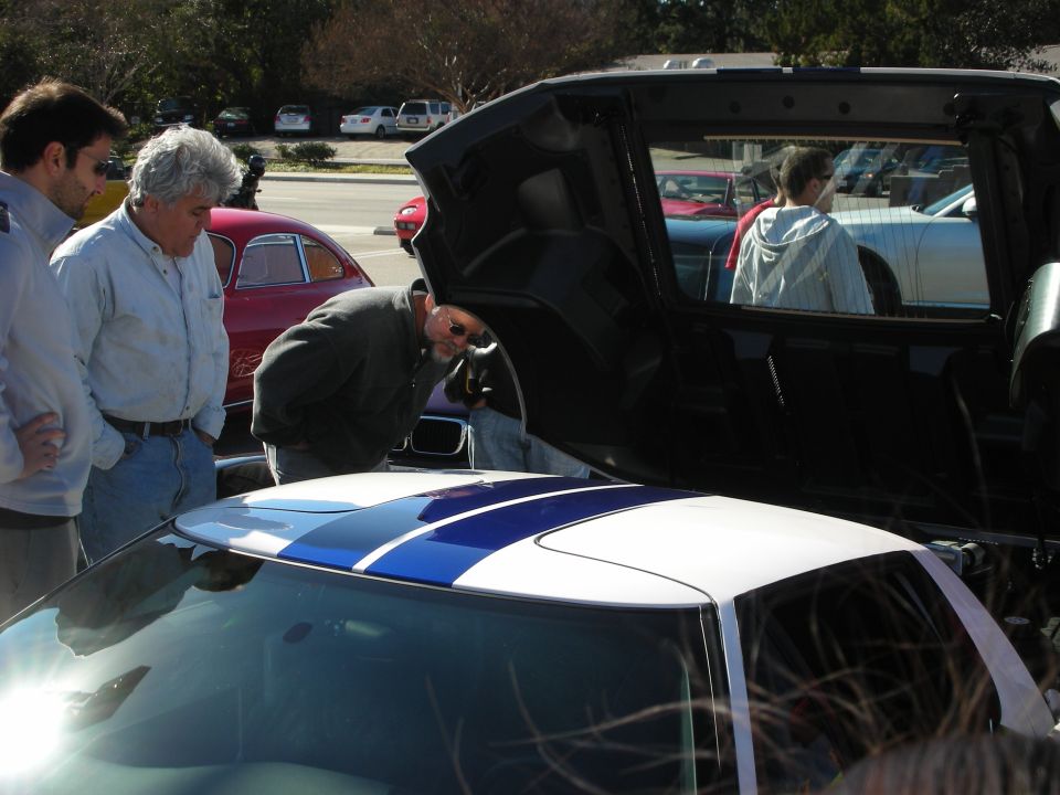 Men examining the engine of a white sports car with blue racing stripes at a parking-lot car meet