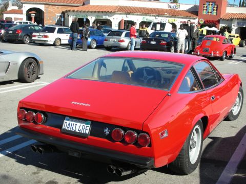 1998 Ford Crown Victoria - Red vintage Ferrari coupe seen from the rear at a car gathering, plate 'DANS 4RE', with other cars and a strip mall behind.