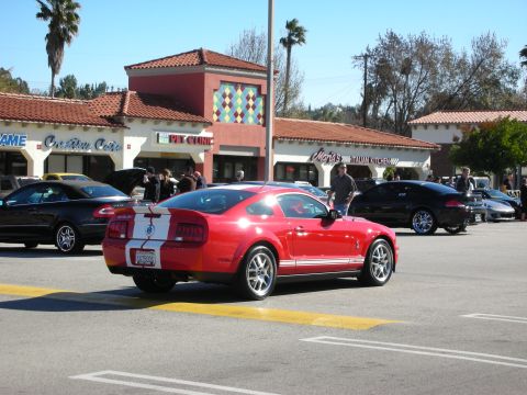1998 Ford Crown Victoria - Red Ford Shelby GT500 Mustang with white racing stripes pulls across a sunlit street lined with parked cars.