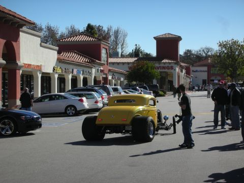 1998 Ford Crown Victoria - Yellow channeled hot rod with exposed front suspension cruises a strip-mall lot as spectators watch nearby.