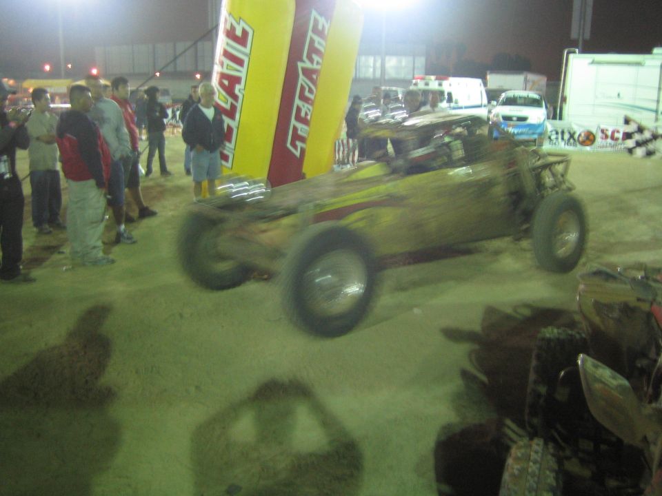 Mud-covered off-road race buggy at night near Tecate arch and crowd