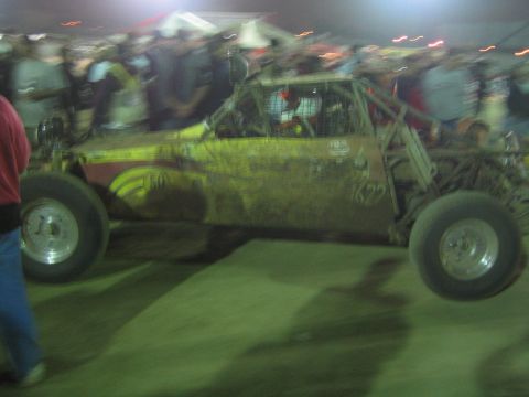 Yellow mud-spattered off-road race car with roll cage in a crowd at night