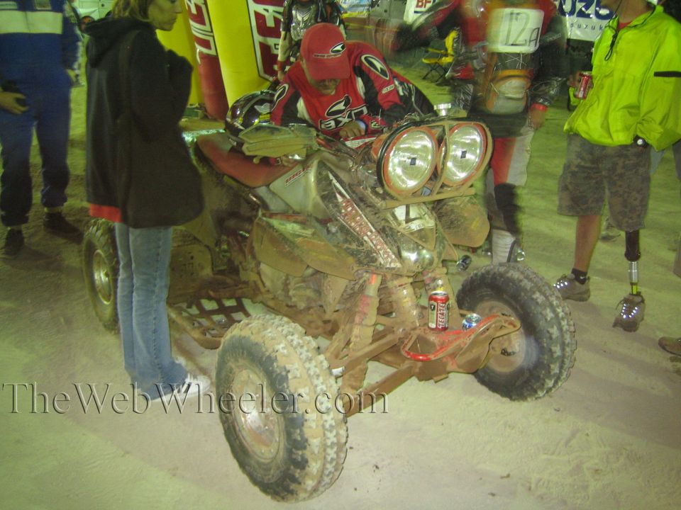 Muddy sport ATV with twin headlights and rider at night, race banners behind