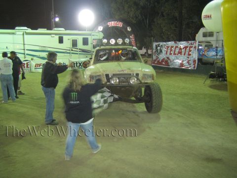 Muddy off-road race truck with light bar at night, person waving checkered flag