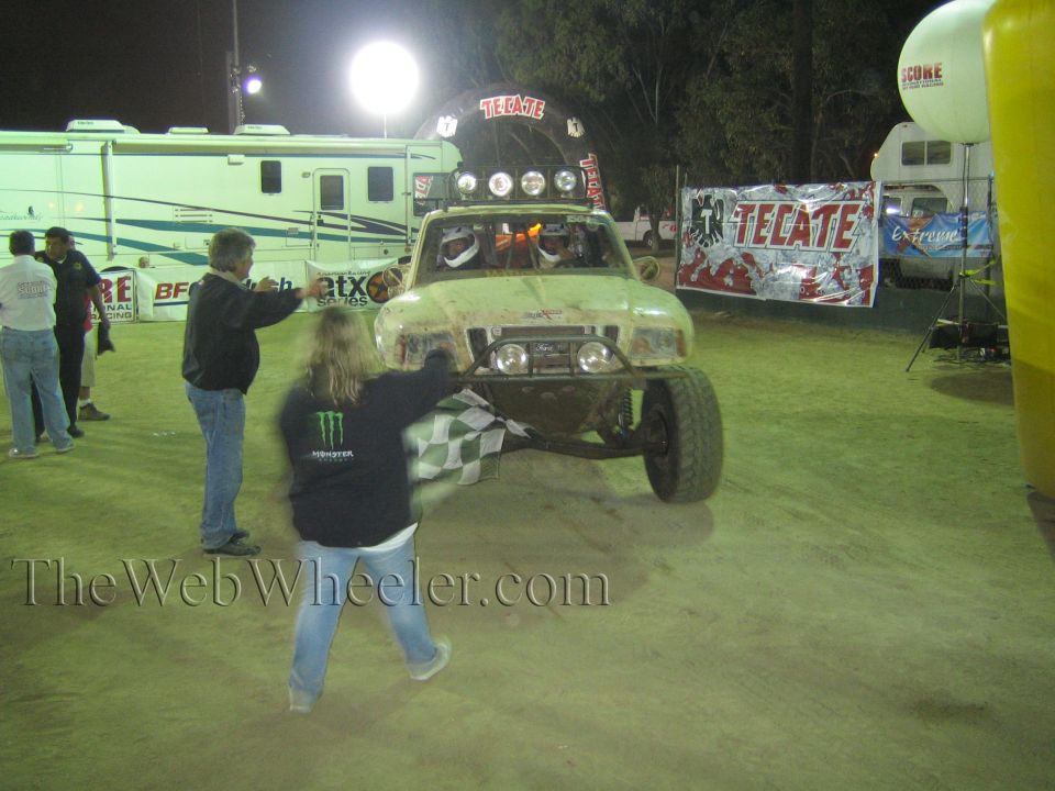 Muddy off-road race truck with light bar at night, person waving checkered flag