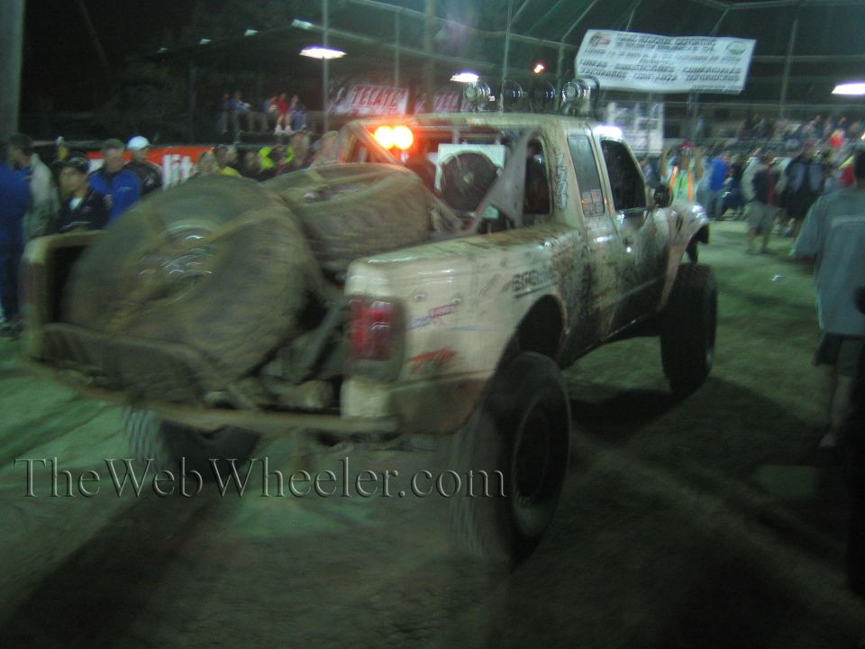 Tan off-road race truck with spare tires speeding at night, crowd behind