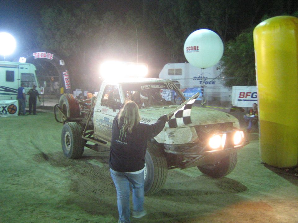 Woman with checkered flag beside muddy off-road race truck under bright lights at night