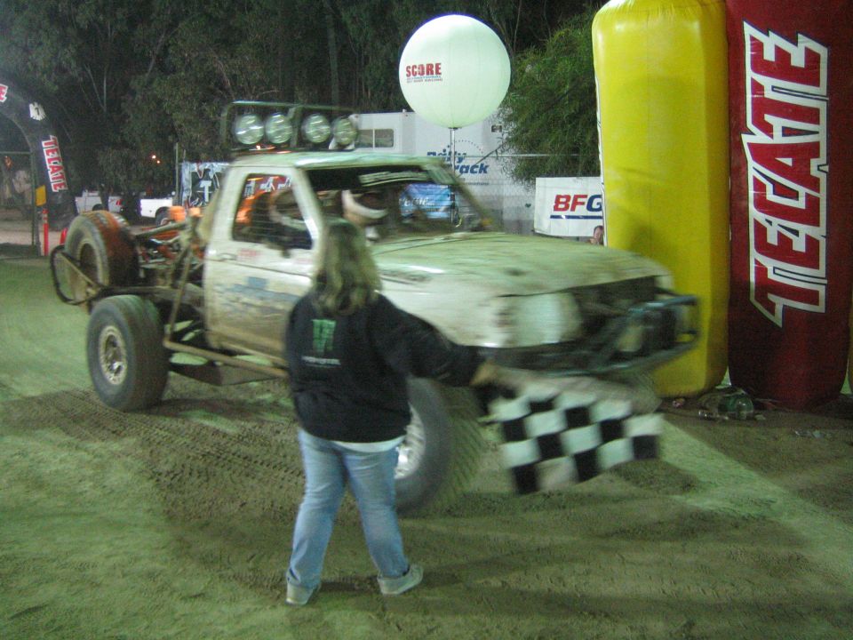 Person waving checkered flag in front of off-road truck near Tecate inflatable arch at night