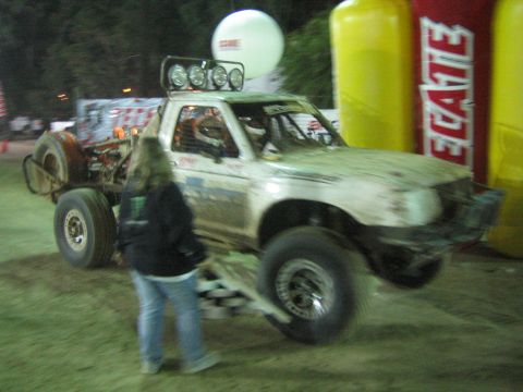 Mud-covered off-road race truck with roof lights creeping past a person at a night event