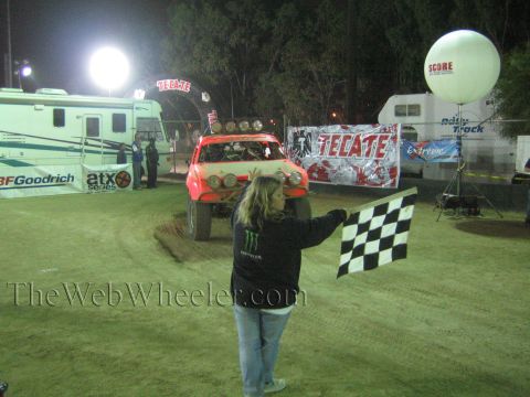 Red off-road race truck at Tecate-banner finish line as a flagger holds a checkered flag