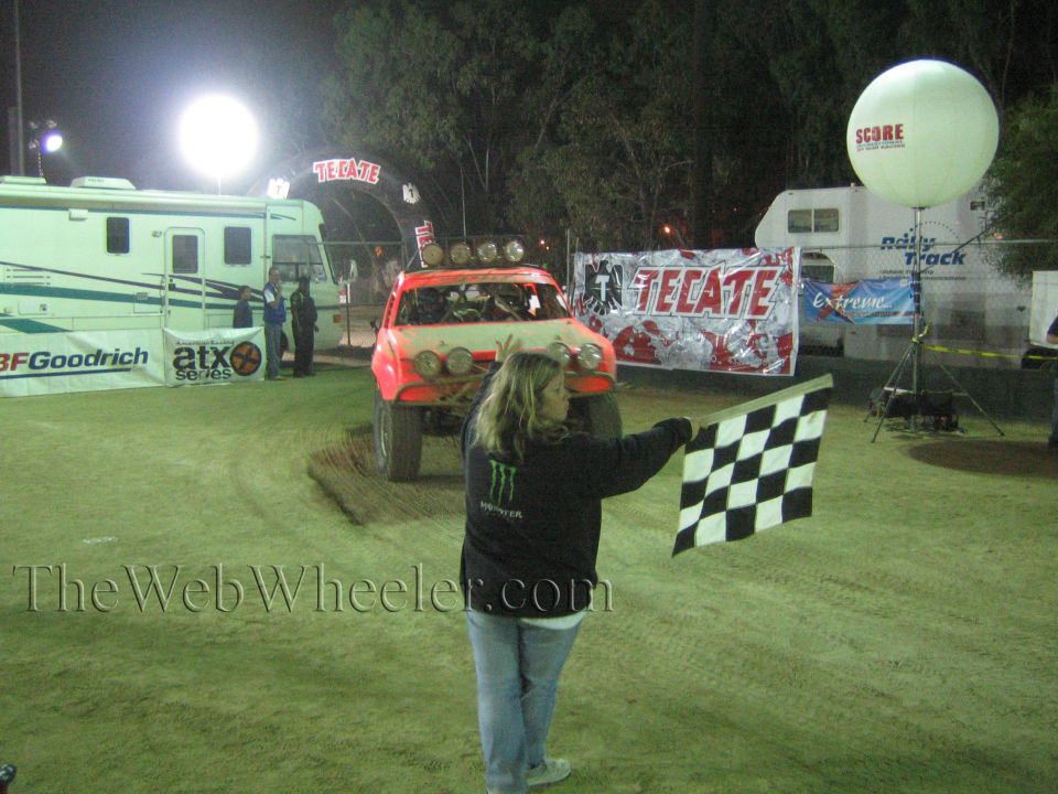 Red off-road race truck at Tecate-banner finish line as a flagger holds a checkered flag