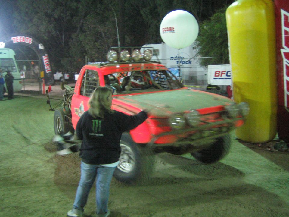 Red off-road race truck with roof lights passing a flagger at a floodlit night race