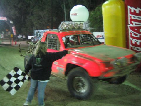Red off-road race truck in motion past a flagger waving a checkered flag at night