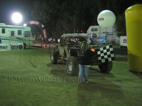 Open-cockpit off-road buggy near Tecate arch as flagger holds checkered flag at night