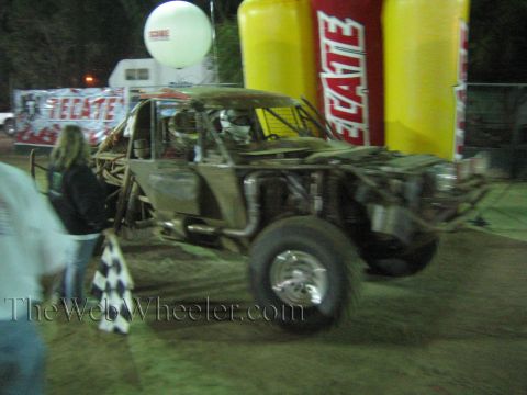 Tube-frame off-road race car with exposed engine beside spectators at a night event
