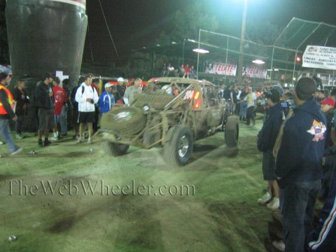 Muddy open off-road buggy surrounded by spectators and a photographer at night