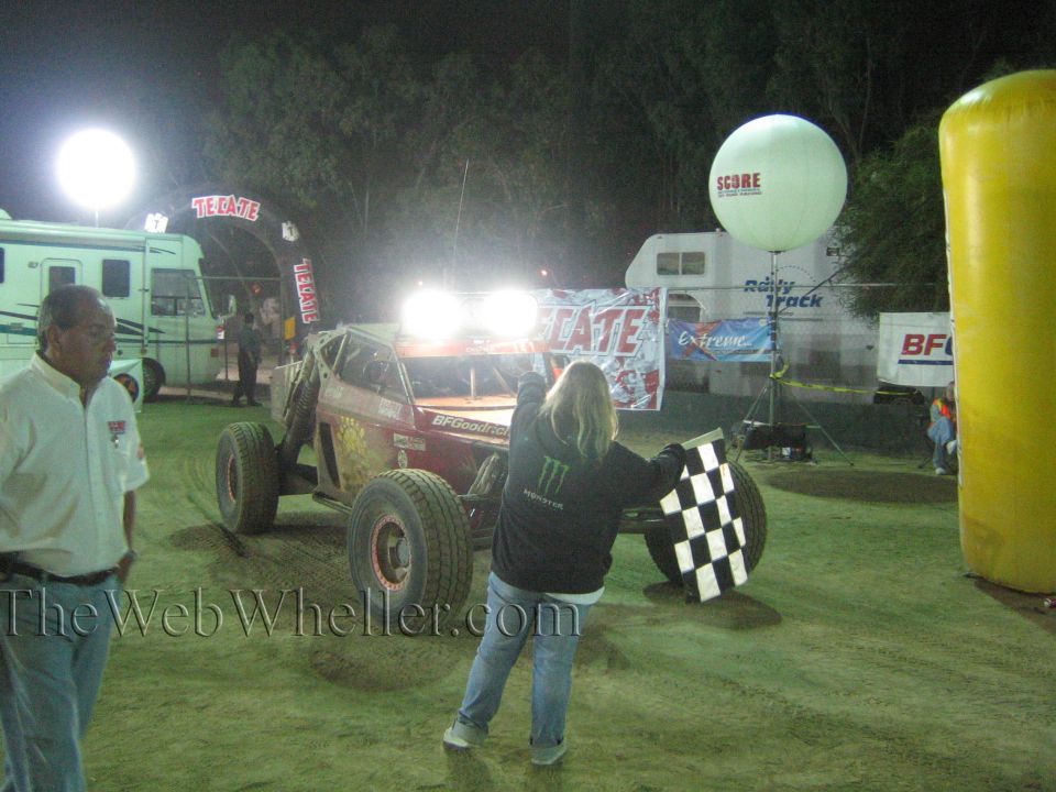 Off-road race car at Tecate finish as a flagger holds a checkered flag under lights