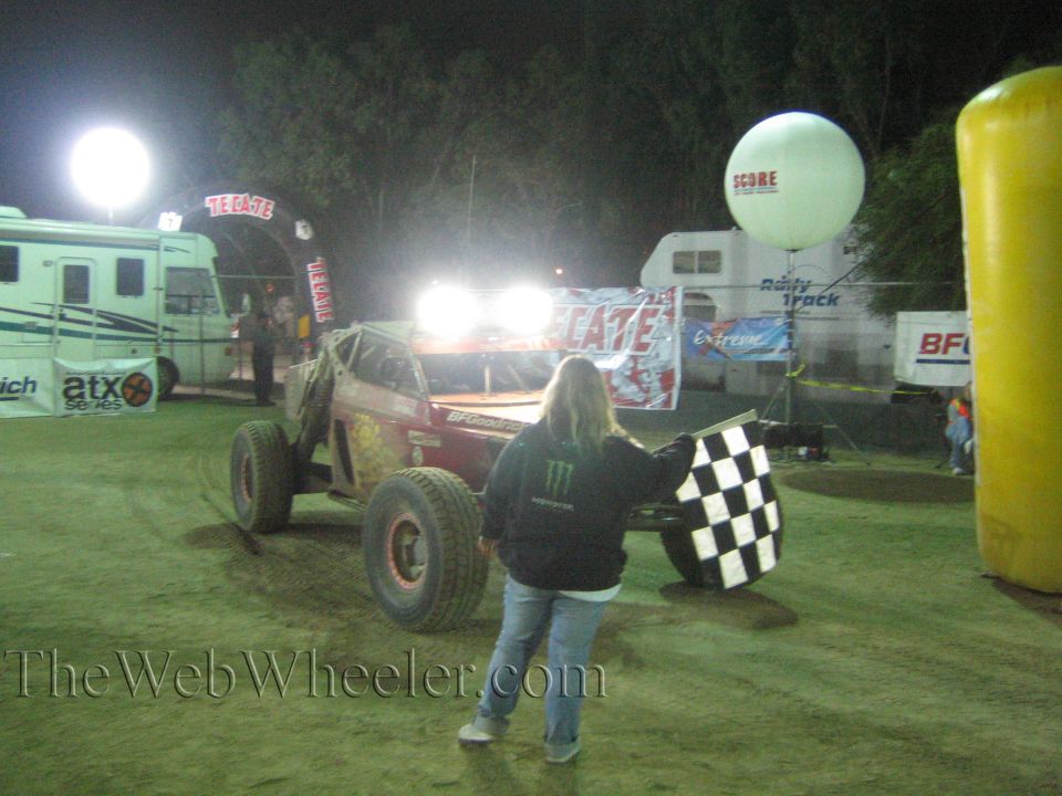 Off-road race car driving toward flagger with checkered flag at a night finish line