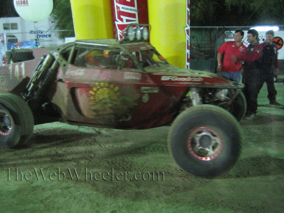 Stickered off-road buggy with sun graphic and onlookers near a Tecate arch at night