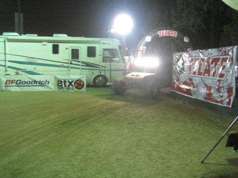 Light-colored off-road buggy parked by an RV and Tecate arch at a night event