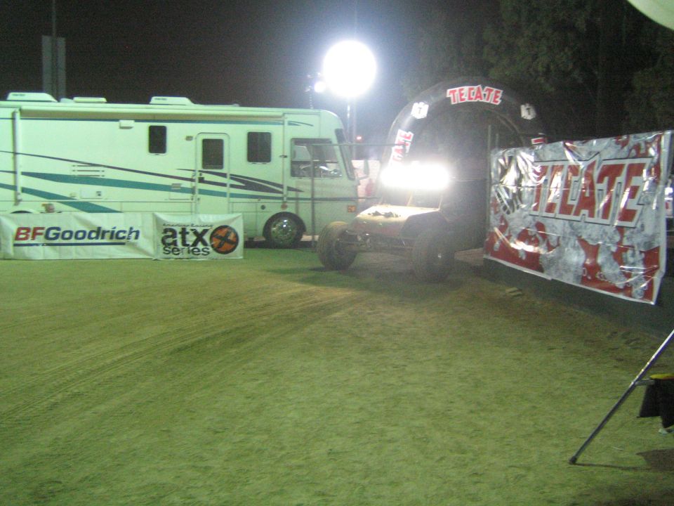Light-colored off-road buggy parked by an RV and Tecate arch at a night event