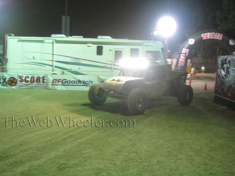 Off-road buggy driving past SCORE and BFGoodrich banners and RV at night