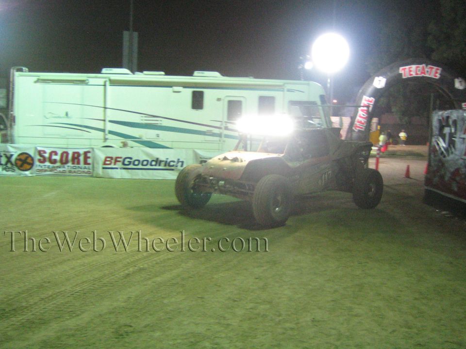 Off-road buggy driving past SCORE and BFGoodrich banners and RV at night