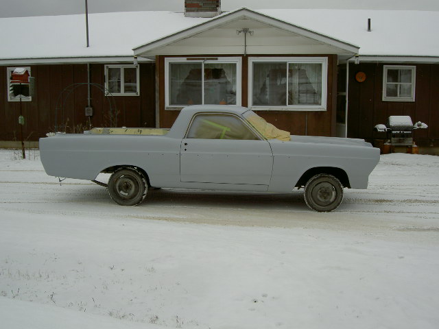 Side view of a 1967 Ford Ranchero in primer, parked in the snow in front of a house
