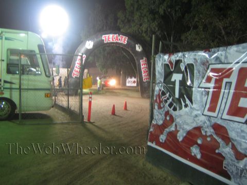 Lit Tecate arch over a dirt course beside a motorhome and banner