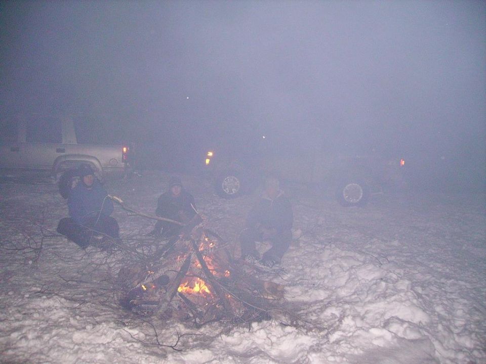 People sit around a campfire in the snow, with two vehicles visible in the hazy background