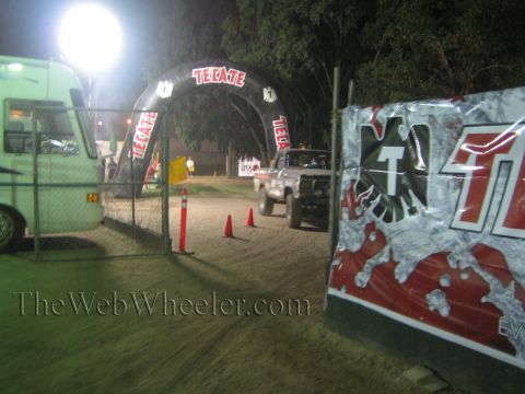 Lifted pickup truck on dirt course under lit Tecate arch at night