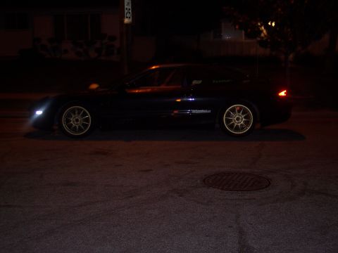 1995 Ford Probe - A dark coupe with gold rims is parked on a street at night, with its headlights illuminated.