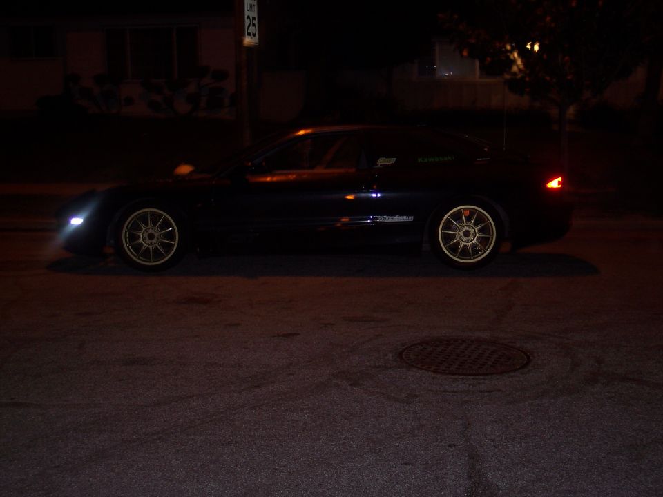 dark coupe parked on street at night, gold rims, headlights on