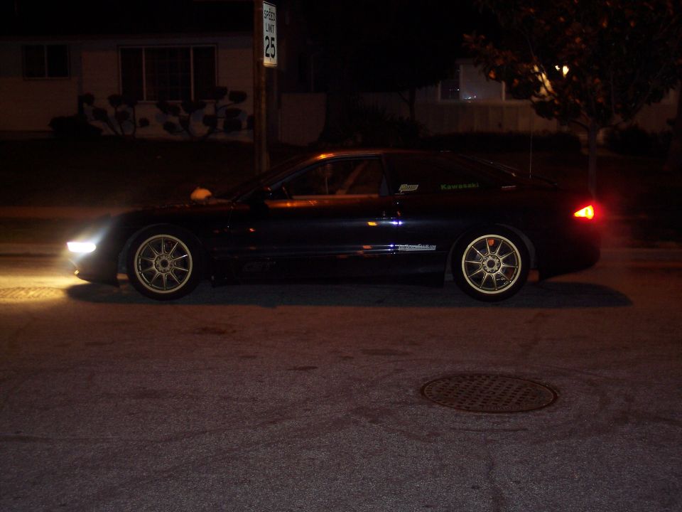 Black coupe with gold multi-spoke wheels parked at night, showing a speed limit sign