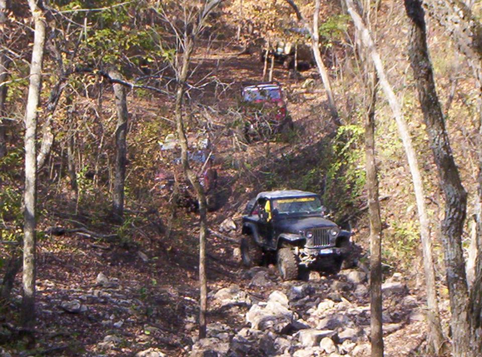 Black lifted Jeep CJ on big tires crawling down a rocky wooded trail, other 4x4s behind
