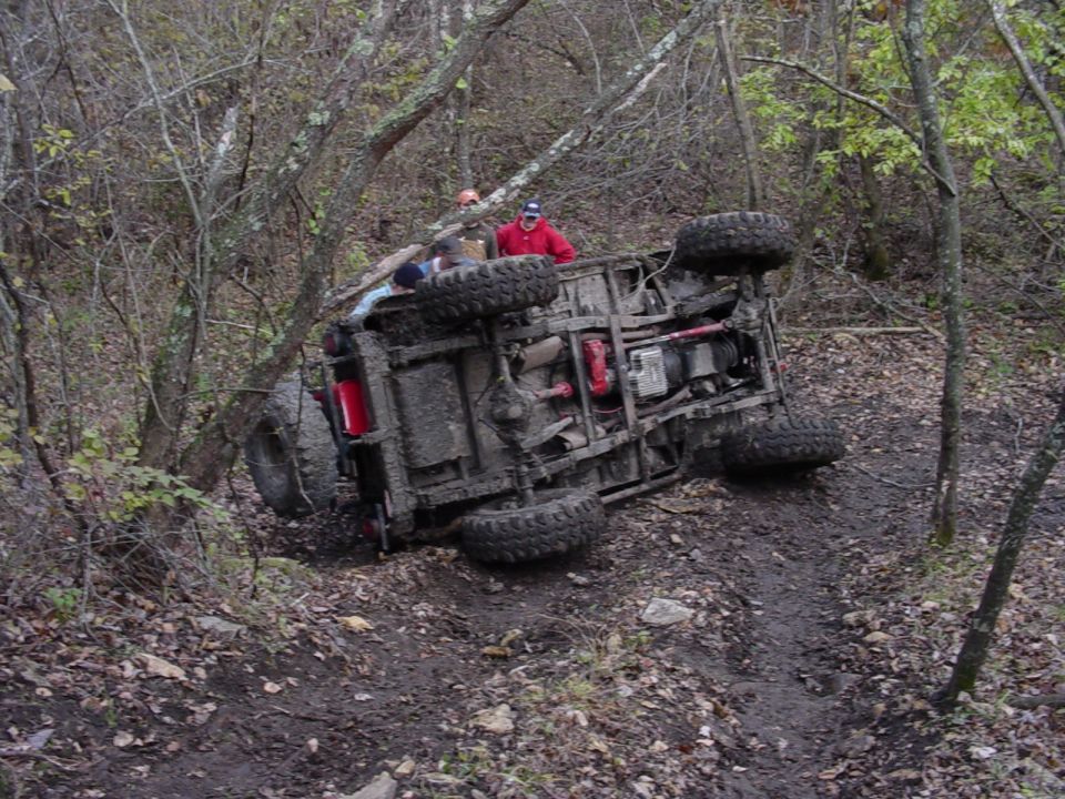 Off-road vehicle rolled on its side on a wooded trail with two onlookers